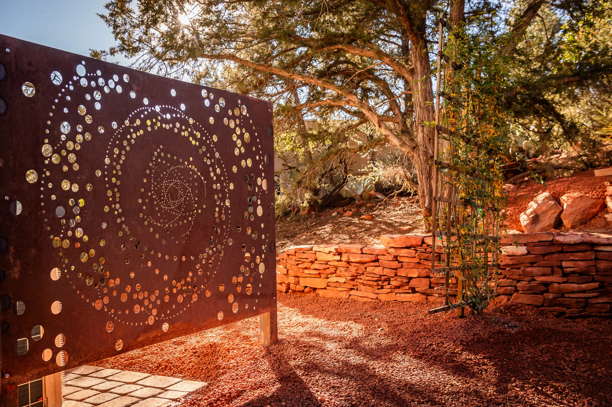 Perforated metal spiral screen in a red rock garden with stacked stone walls and lush juniper trees.