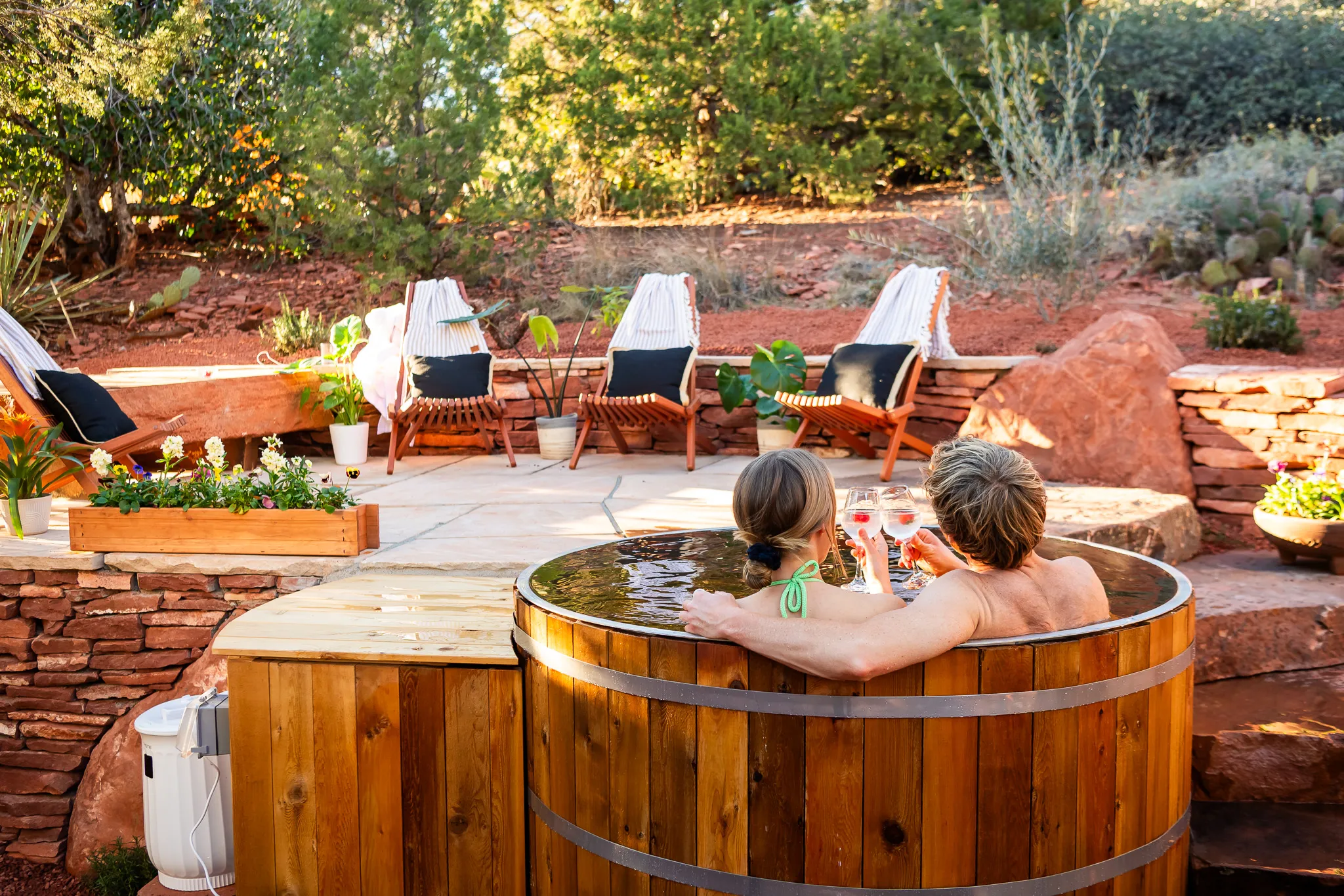Couple toasts with drinks in a cedar soaking tub on a stone patio surrounded by Sedona's red rocks.