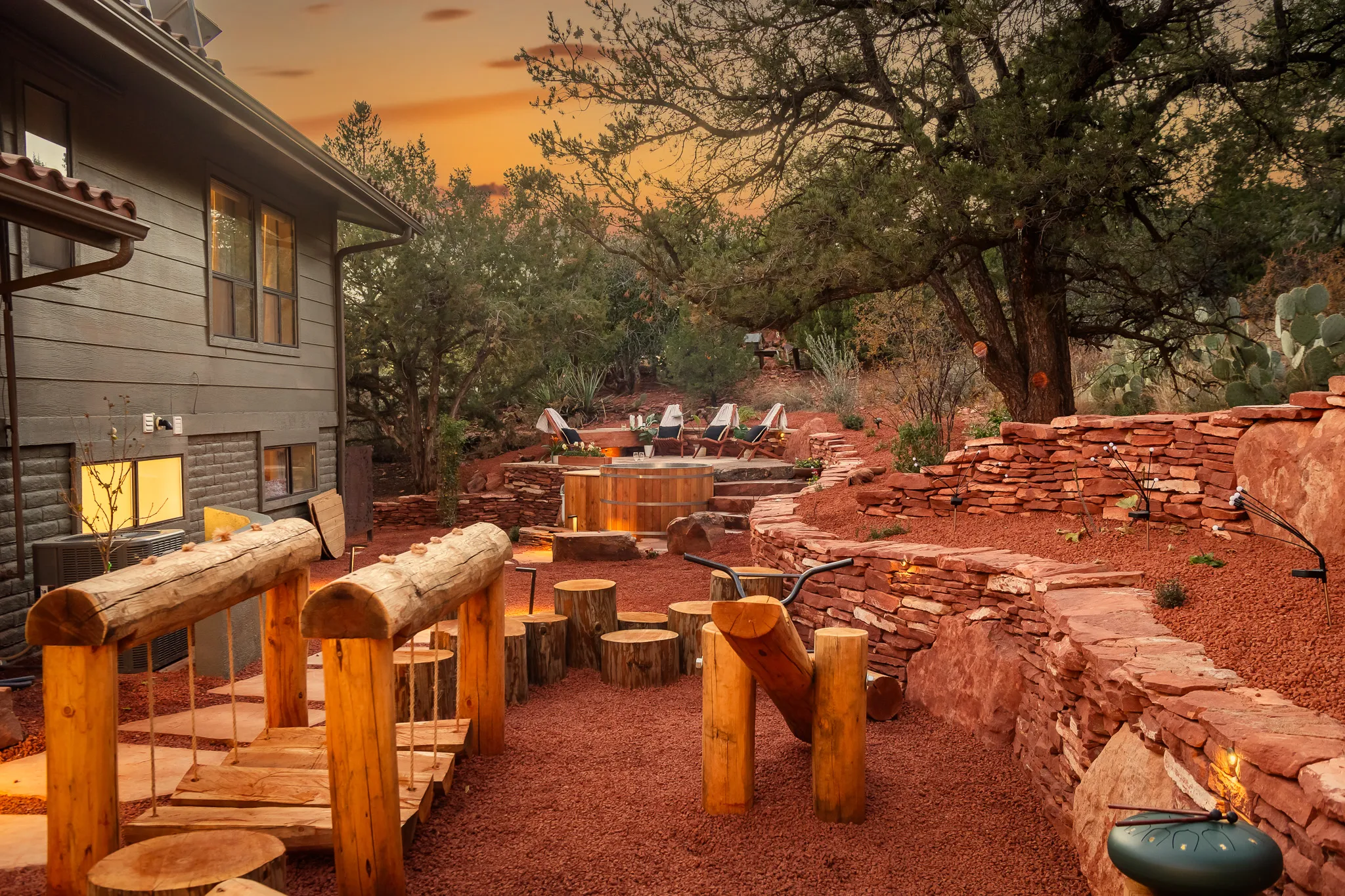 Wooden log playground bridge and stumps on red gravel with a cedar hot tub and stone walls under a sunset sky.