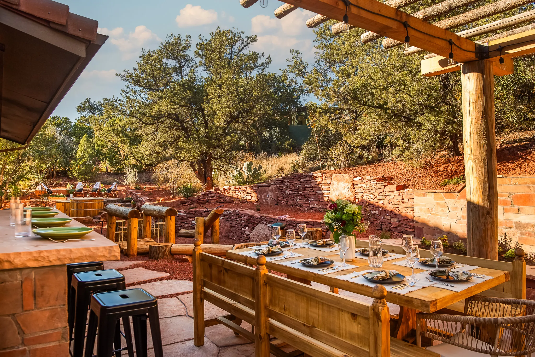 Wooden dining table under a pergola and outdoor kitchen bar set against a lush Sedona landscape.