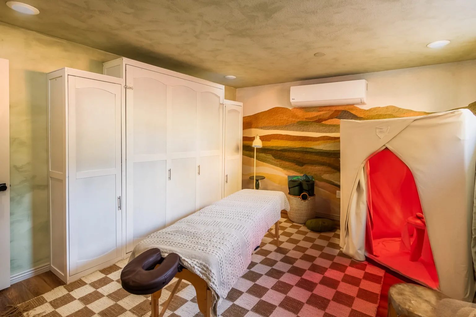 Massage table and portable infrared sauna in a wellness room with a mountain landscape mural.